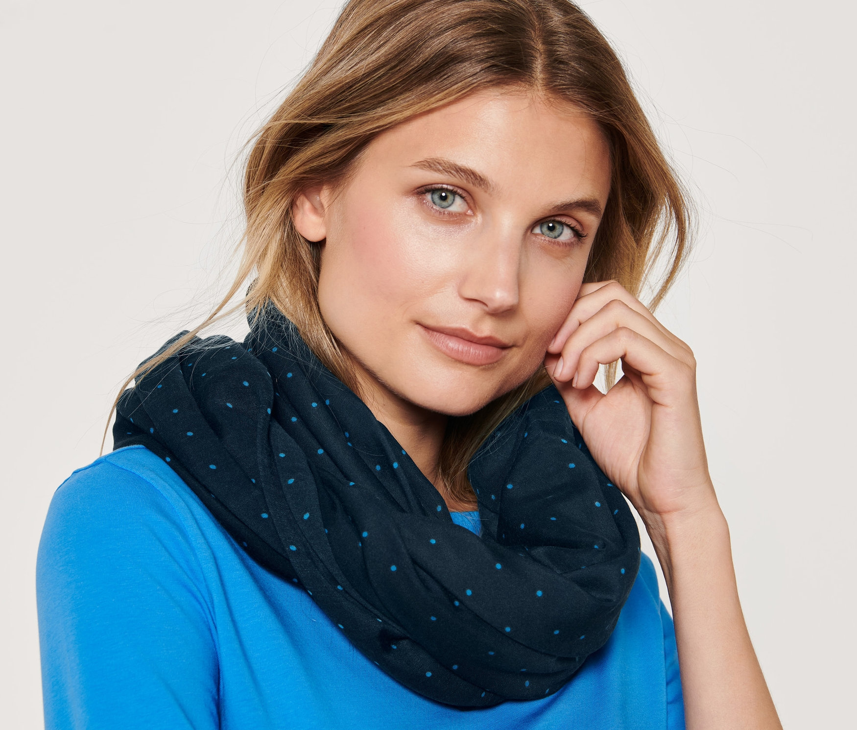 Portret blondwłosej kobiety o niebieskich oczach, ubranej w szal typu komin w kropki i niebieską koszulkę.