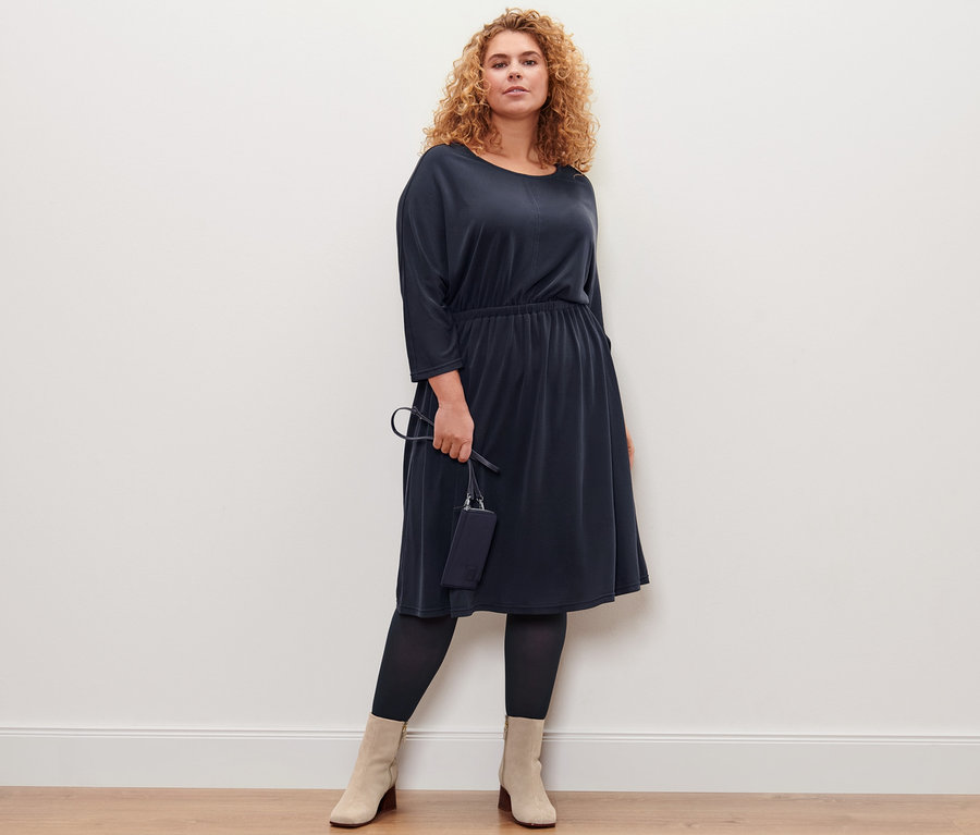 Kobieta pozuje w sukience dżersejowej o splocie piqué i trzyma minitorebkę. Ma na sobie cienkie rajstopy, 2 pary.