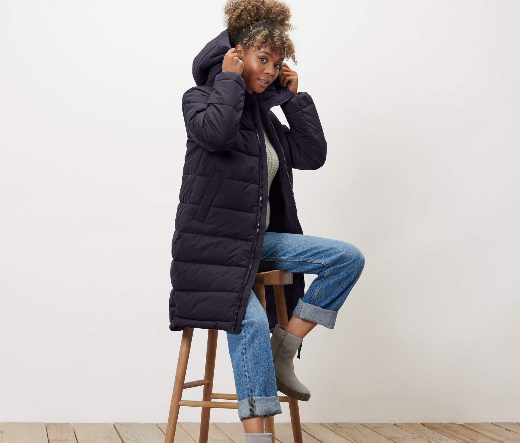 Kobieta siedzi na stołku i poprawia kaptur niebieskiego pikowanego płaszcza.