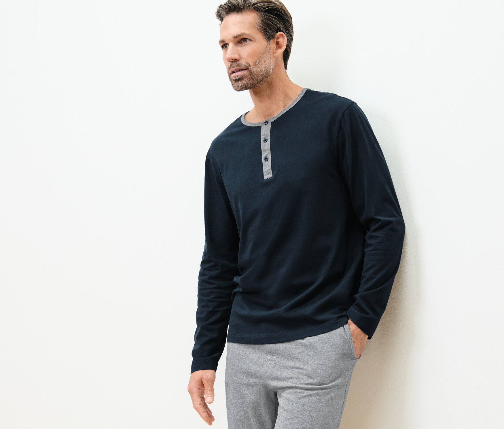 Mężczyzna oparty o ścianę ma na sobie koszulki od piżamy (2 sztuki) i szare spodnie.