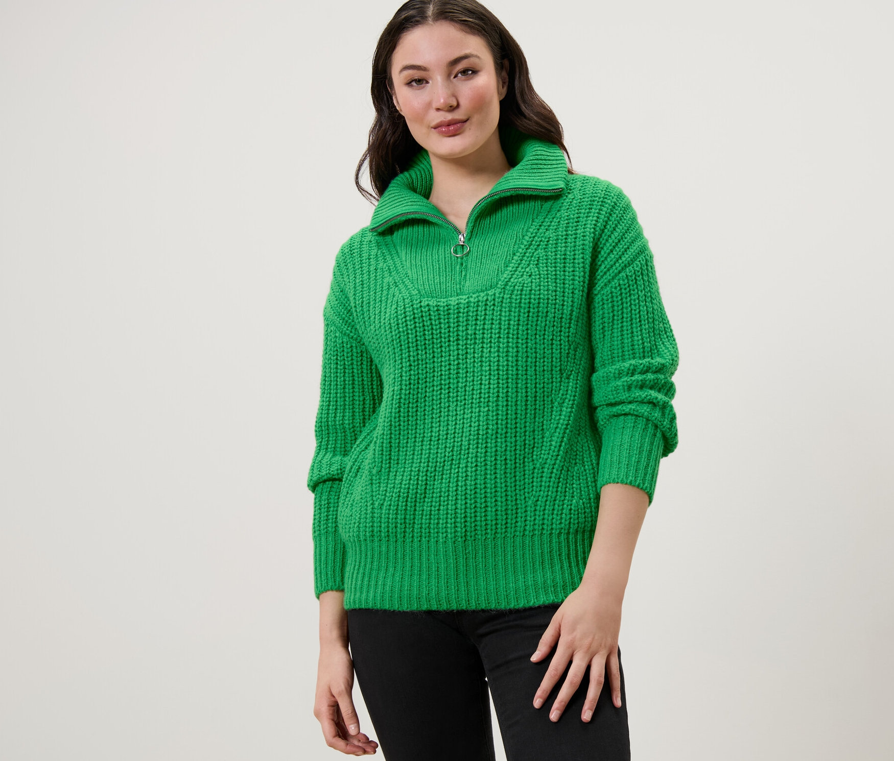 Kobieta ma na sobie zielony sweter o grubym splocie z kołnierzem typu troyer i czarne dżinsy.