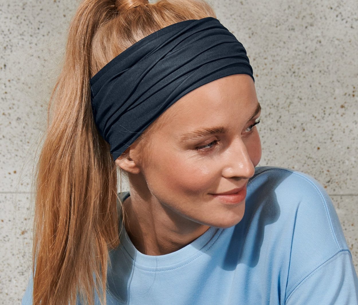 Kobieta z blond włosami związanymi w kucyk nosi chustkę wielofunkcyjną i niebieski top.