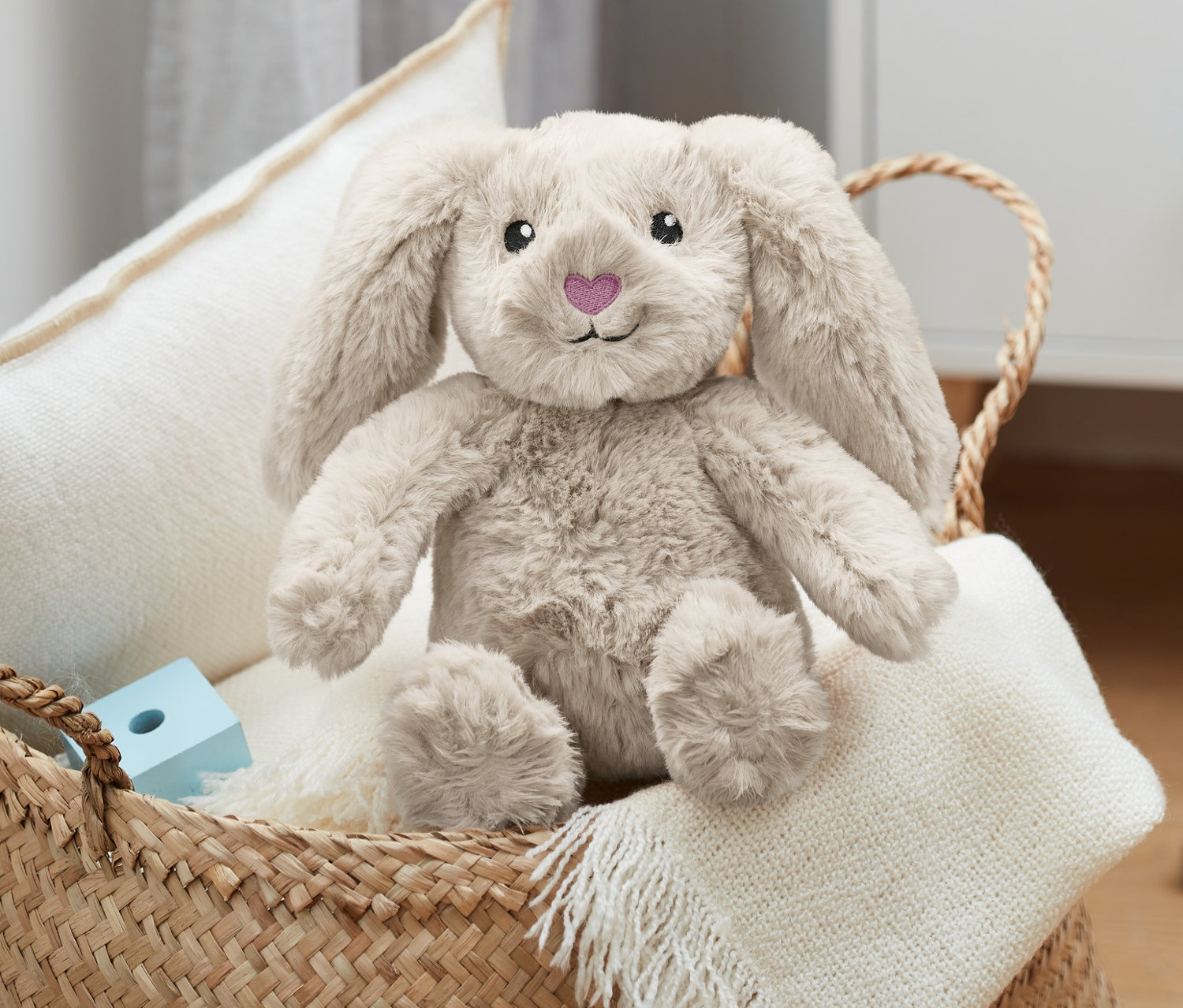 Pluszowy zając siedzi w wiklinowym koszu z białą poduszką i kocem.