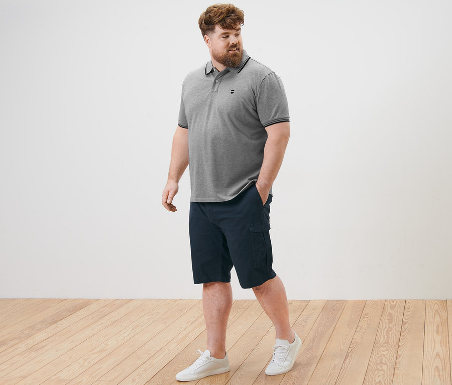 Mężczyzna idzie po drewnianej podłodze, ubrany w szarą koszulkę polo o splocie pique i granatowe szorty bojówki.