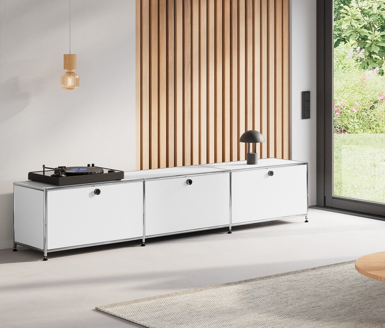 Pokój dzienny z okrągłym stolikiem kawowym (80 cm), metalową niską szafką »CN3« z 3 białymi przegródkami z klapami, dodatkową pomarańczową klapą metalową „CN3” i drewnianą lampą wiszącą.