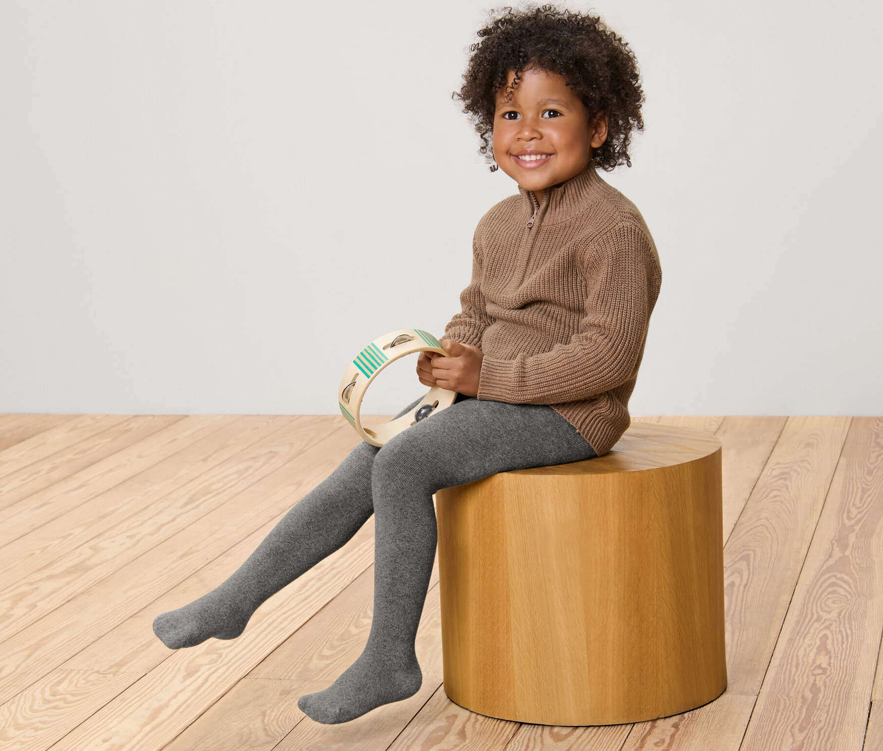 Uśmiechnięte dziecko siedzi na drewnianym stołku i trzyma tamburyn. Ma na sobie brązowy sweter i szare, melanżowe dziecięce rajstopy termoaktywne, 2 pary, melanż.