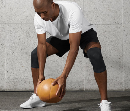 Mężczyzna w białej koszulce i czarnych szortach trzyma piłkę lekarską i nosi opaski na kolana do fitnessu, 2 sztuki.