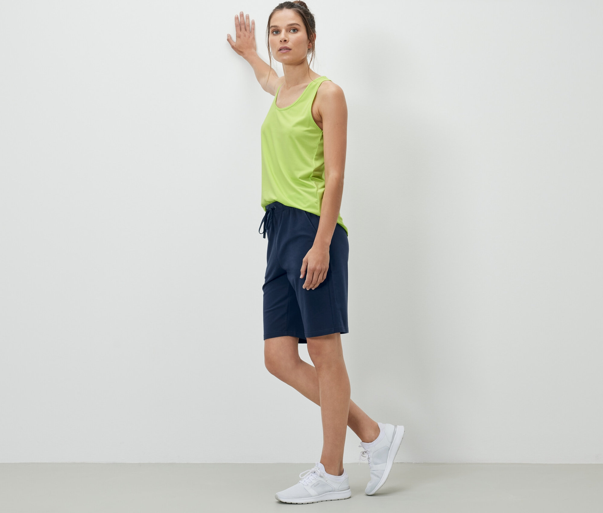 Kobieta oparta o ścianę ma na sobie granatowe szorty dresowe.