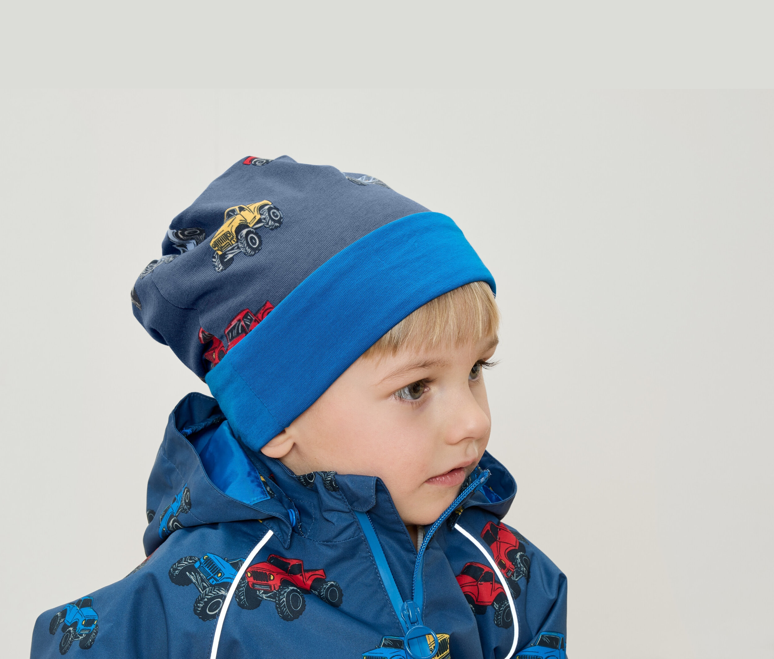 Chłopiec o blond włosach ma na sobie niebieską czapkę i kurtkę z nadrukiem monster trucków.