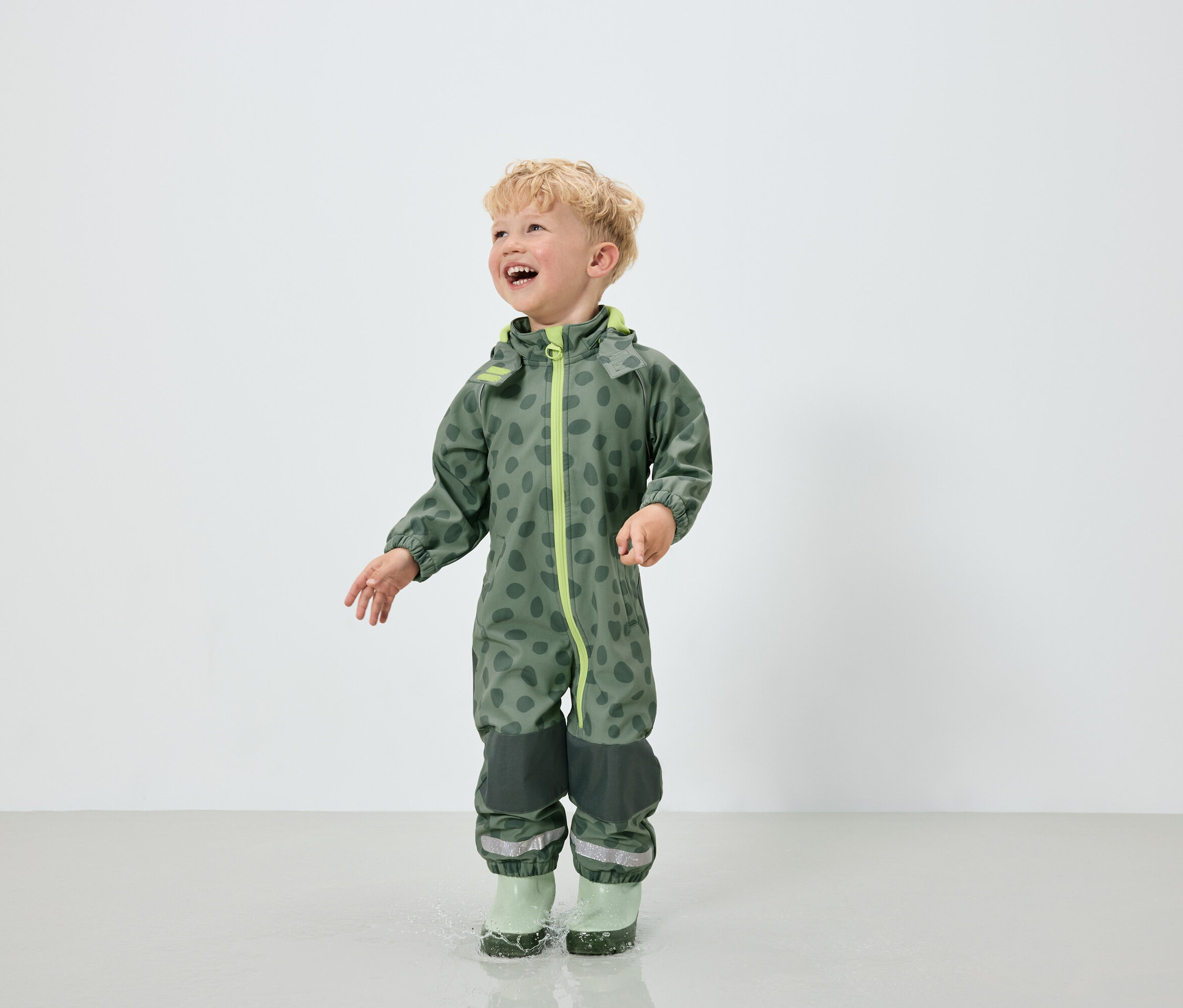 Chłopiec w zielonym kombinezonie we wzór w plamy i zielonych kaloszach skacze w kałuży.