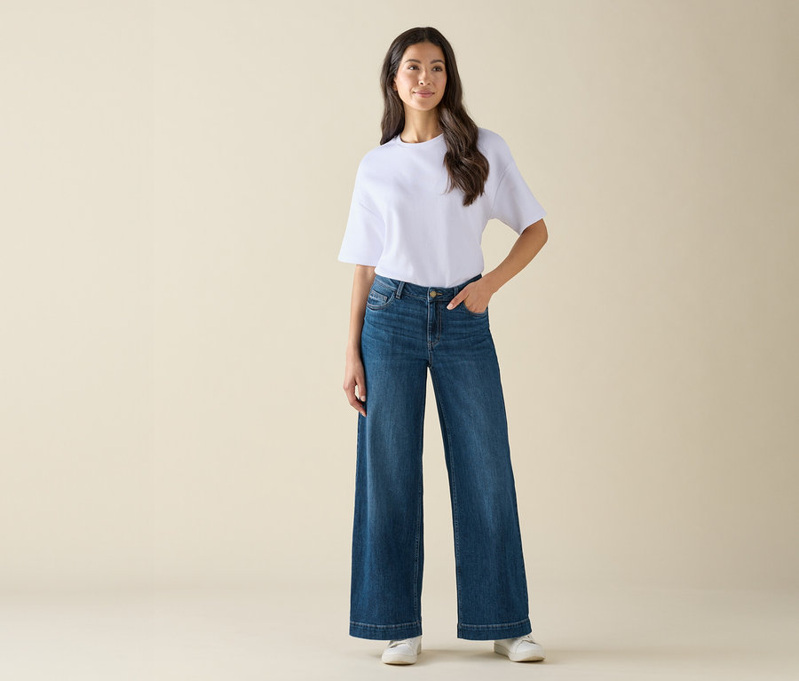Kobieta w białej koszulce i szerokich niebieskich dżinsach pozuje na jasnym tle.