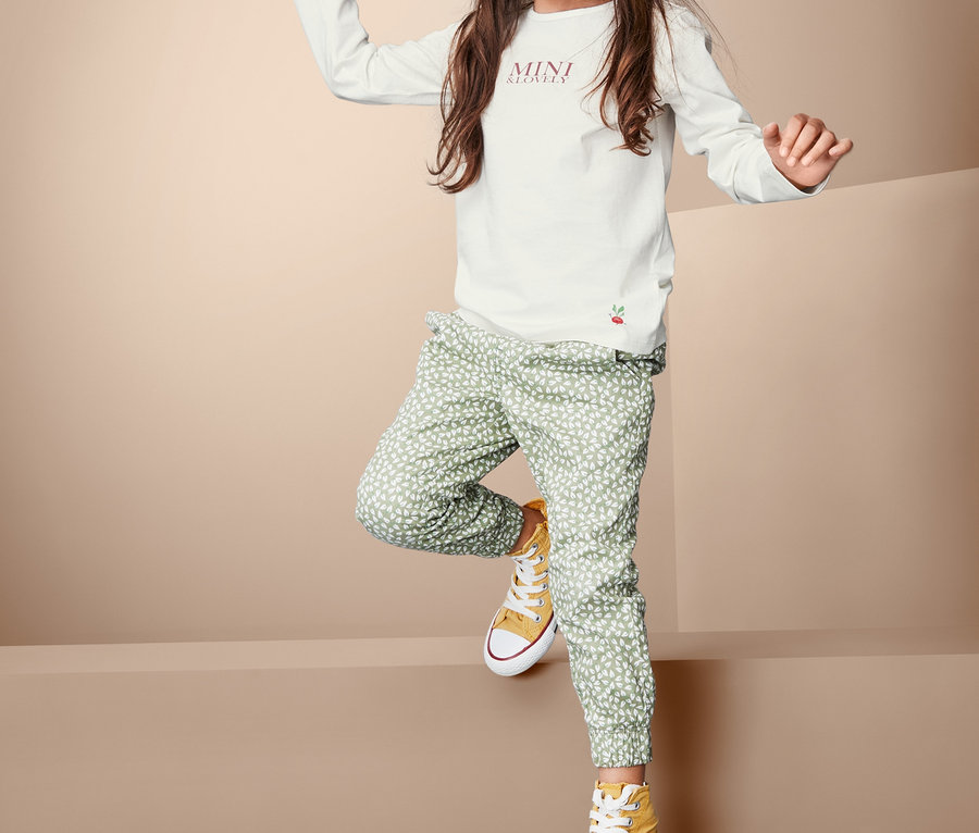 Dziewczynka pozuje w białej koszulce z długim rękawem, zielonych wzorzystych spodniach i żółtych trampkach.