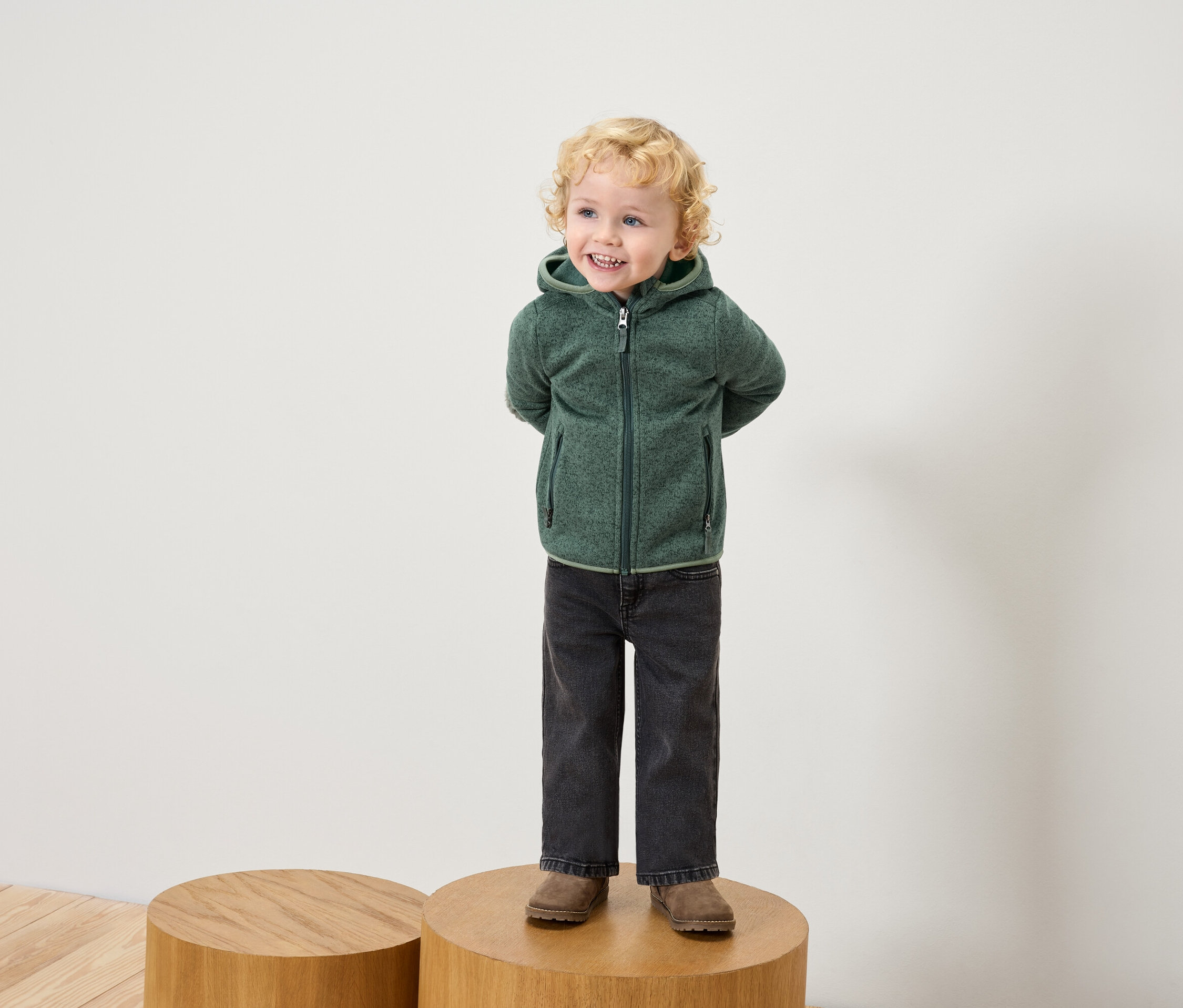 Chłopiec z blond kręconymi włosami stoi na drewnianym cokole, ubrany w zieloną bluzę polarową z kapturem, czarne dżinsy i brązowe buty.