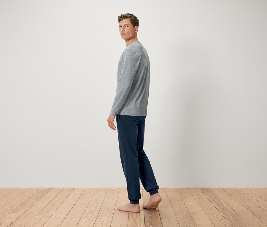 Mężczyzna w szarej koszulce z długim rękawem i granatowych spodniach stoi boso na drewnianej podłodze i patrzy przez ramię.