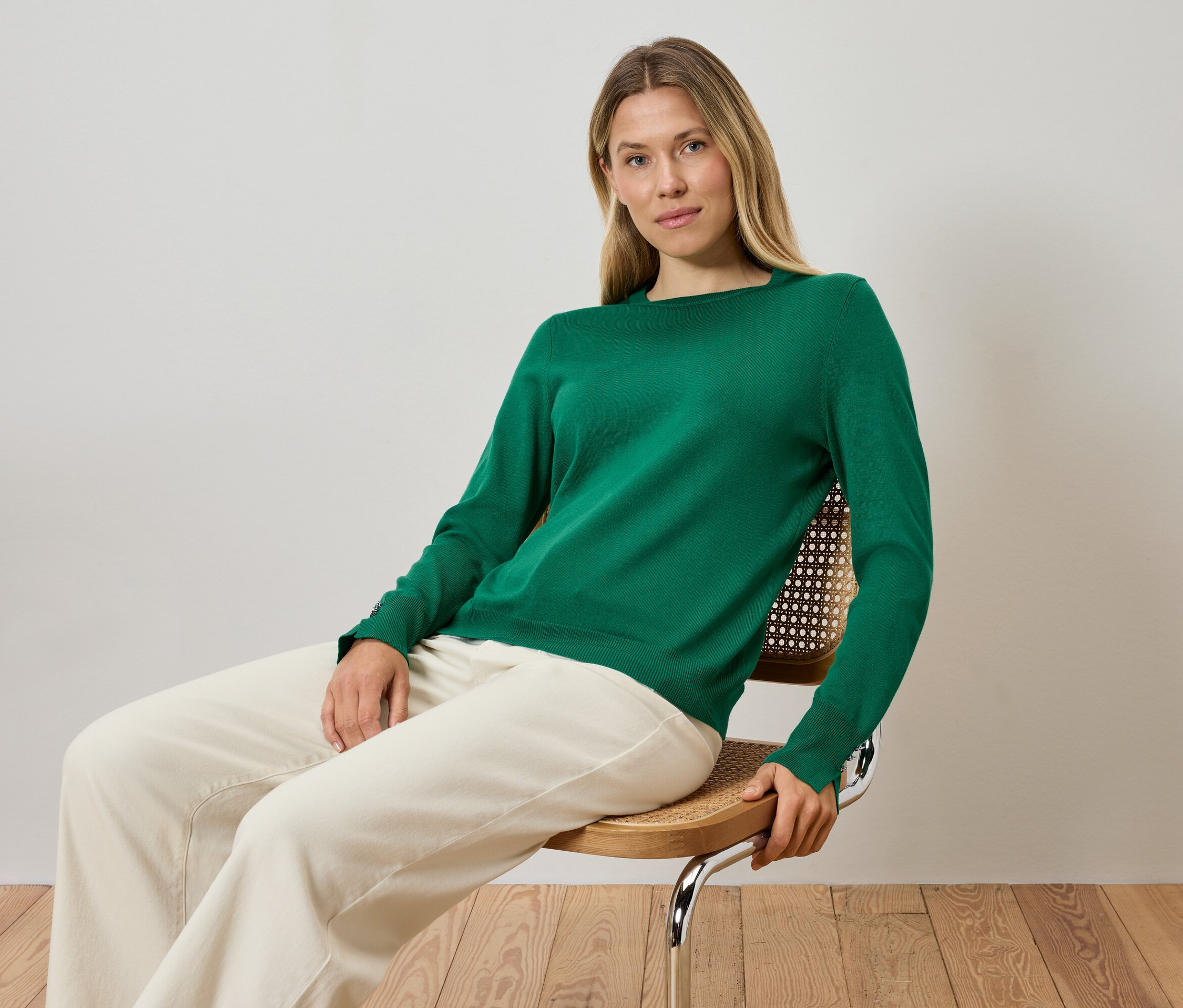 Blondynka siedzi na krześle, ubrana w zielony sweter i kremowe spodnie.