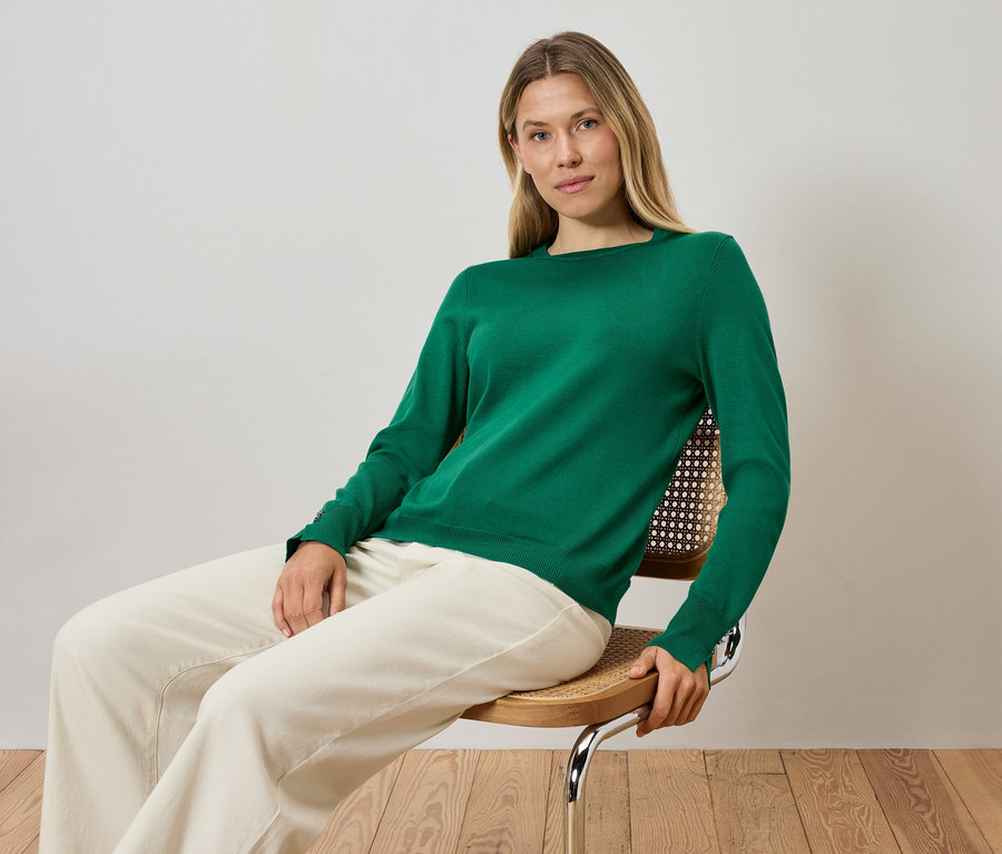 Blondynka siedzi na krześle, ubrana w zielony sweter i kremowe spodnie.