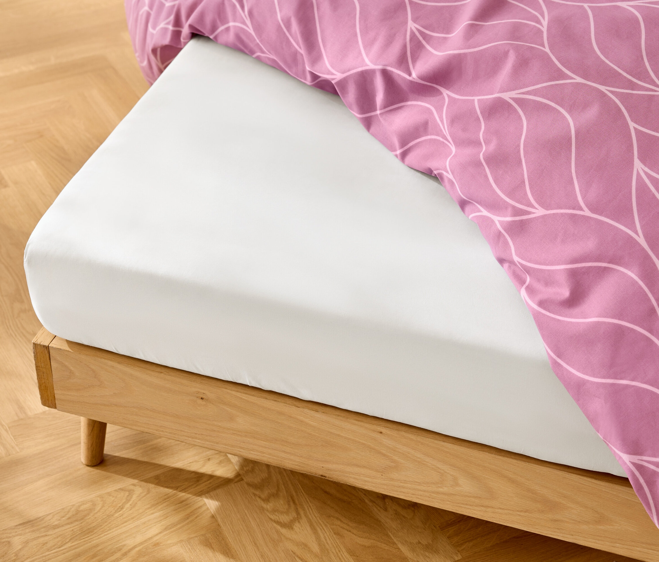 Białe prześcieradło z gumką na drewnianej ramie łóżka z różową pościelą w biały wzór.