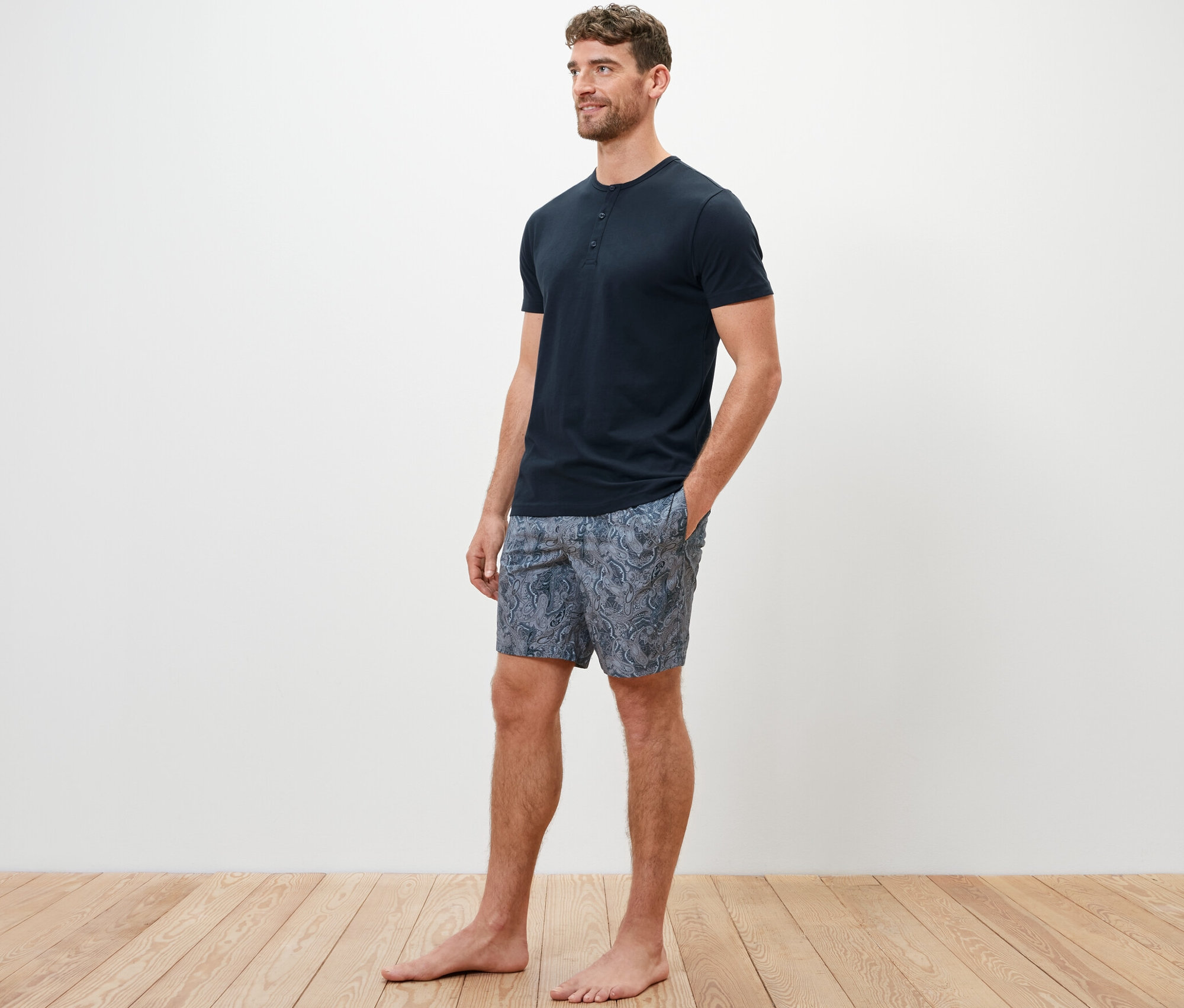 Mężczyzna w piżamie z krótkimi spodenkami z tkaniny stoi boso na drewnianej podłodze.