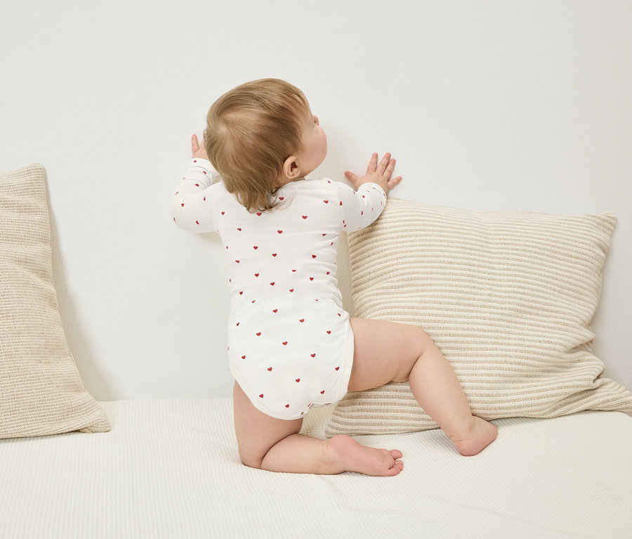 Niemowlę w body z długim rękawem w czerwone serduszka stoi przy ścianie obok poduszki w paski.