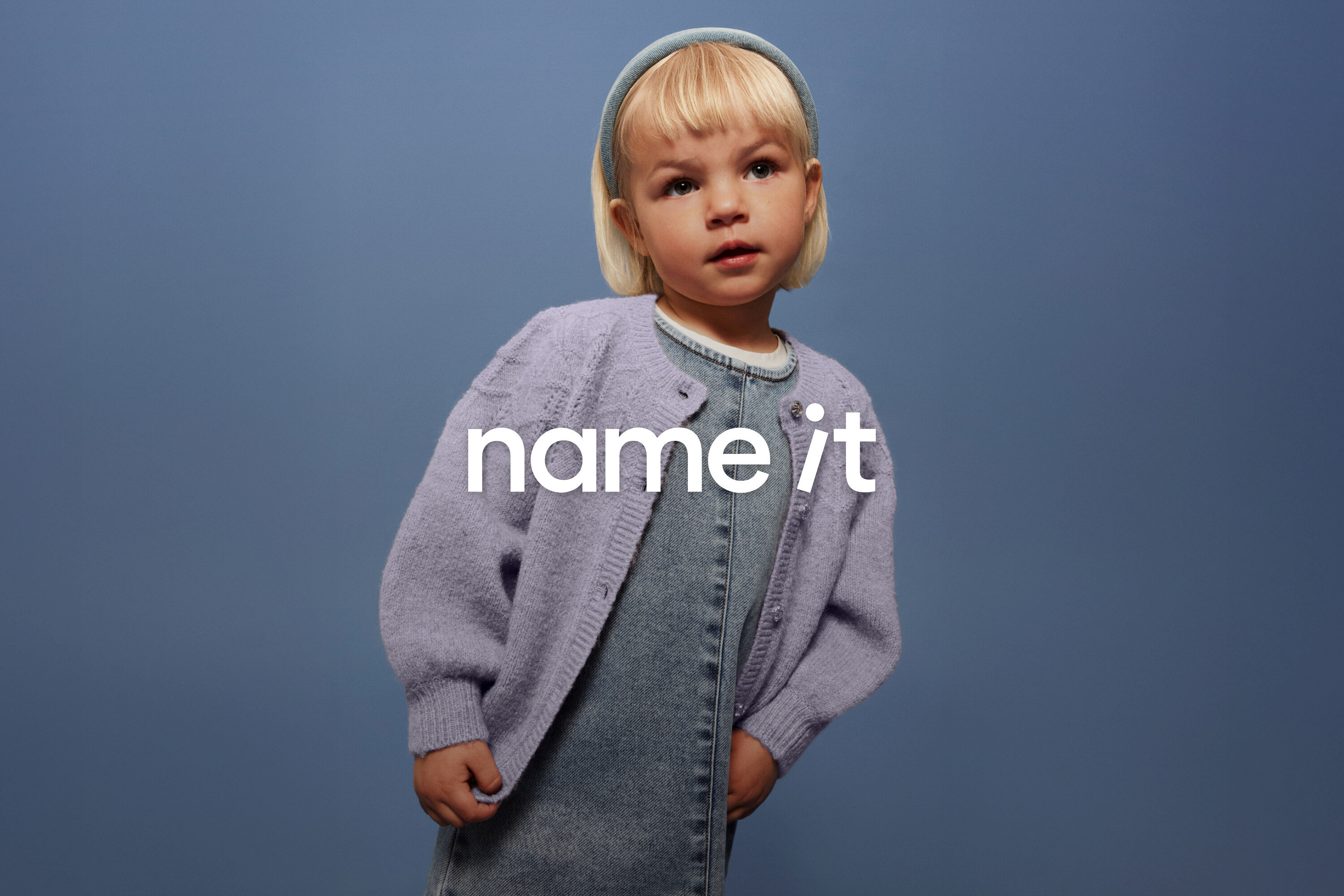 Małe dziecko z blond włosami i niebieską opaską na głowie ma na sobie dżinsowe ogrodniczki i liliowy kardigan na tle niebieskiego tła.