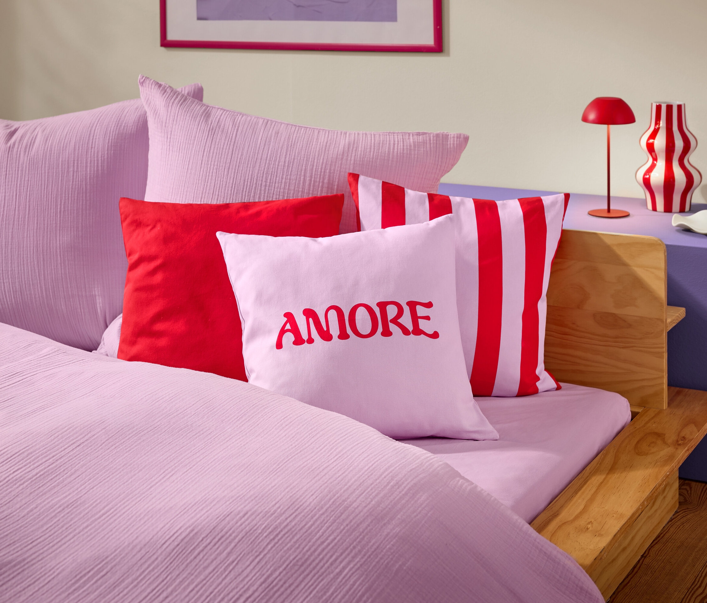 Zbliżenie łóżka z różową pościelą i poduszkami. Na jednej z poduszek widnieje słowo AMORE napisanym na czerwono.