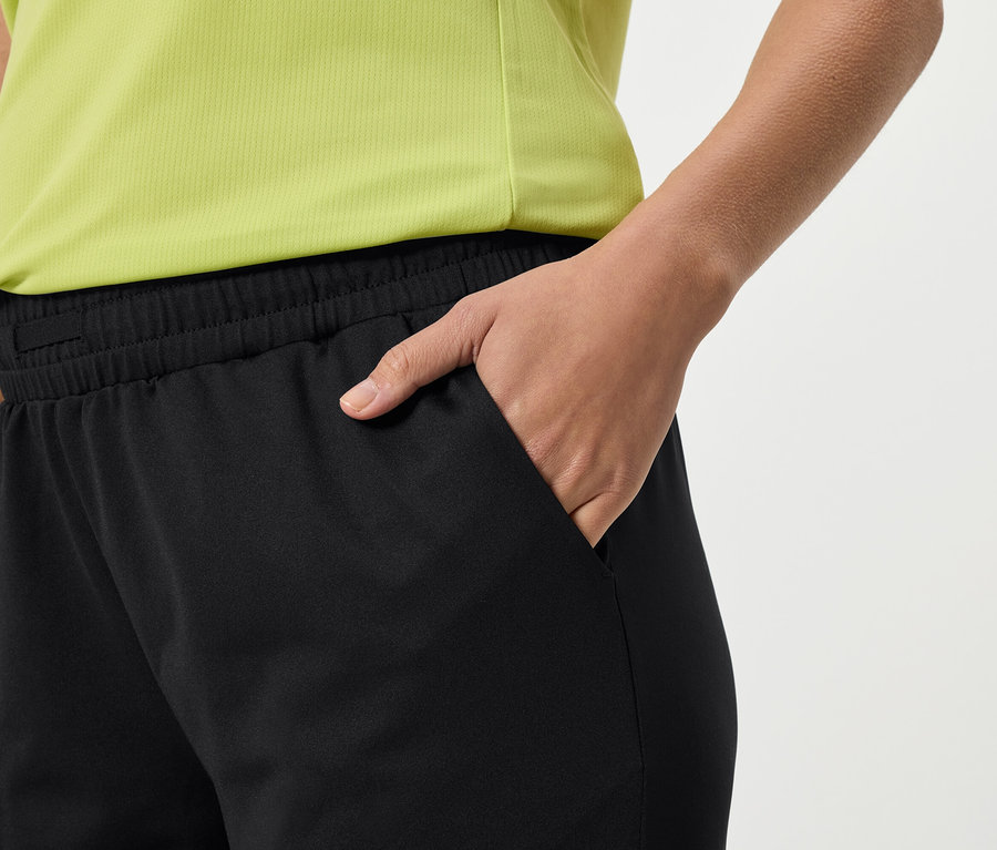 Zbliżenie osoby w czarnych spodniach dresowych i żółtej koszulce, wkładającej rękę do kieszeni.