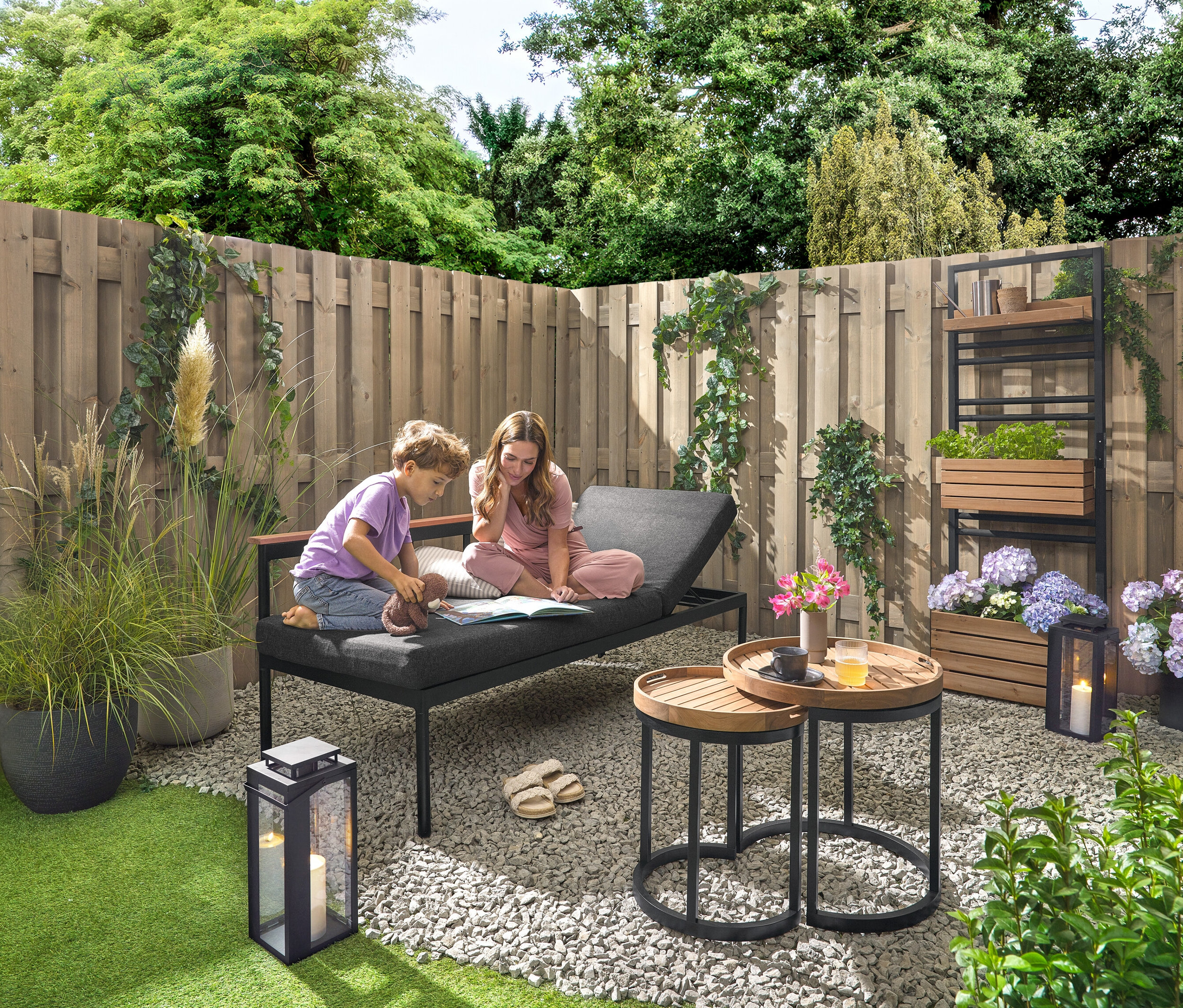 Dziecko i kobieta siedzą na ławce ogrodowej 2 w 1 ‒ leżance »Elin« i czytają książkę. W tle znajduje się regał z donicami i z półką do indywidualnego rozmieszczenia „Elin”. Po prawej stronie stoi lampion ogrodowy ze świecą. Dwa stoliki ogrodowe z odłączanymi tackami, Ø ok. 40 cm i 50 cm, oraz 2 poduszki ogrodowe z lnu.