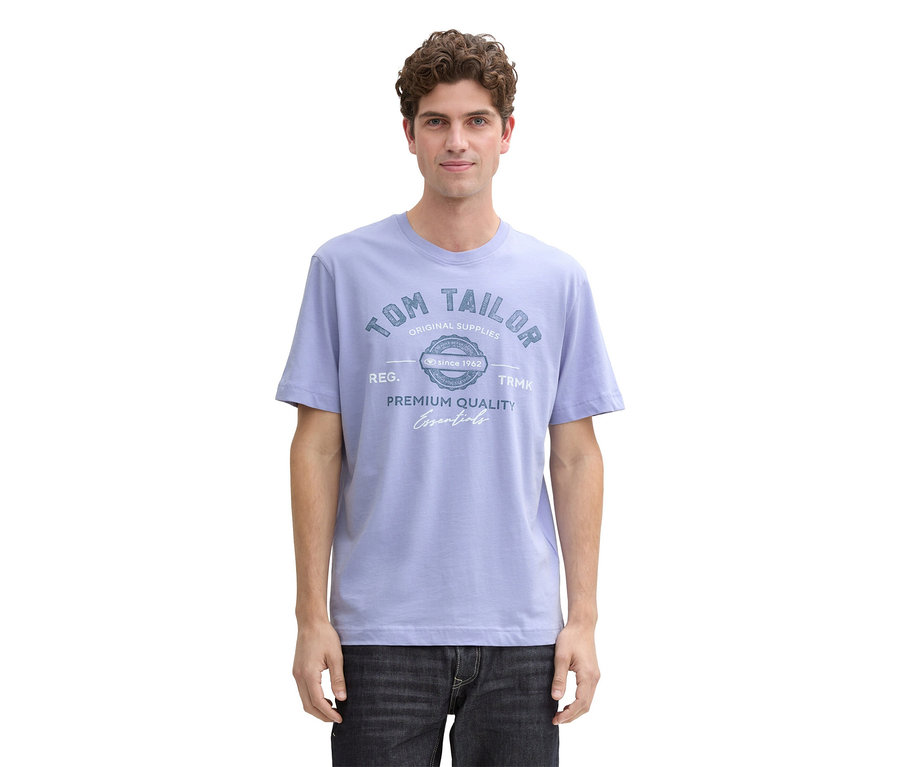 Mężczyzna o kręconych włosach ma na sobie jasnoniebieski T-shirt z logo Tom Tailor i ciemne dżinsy.