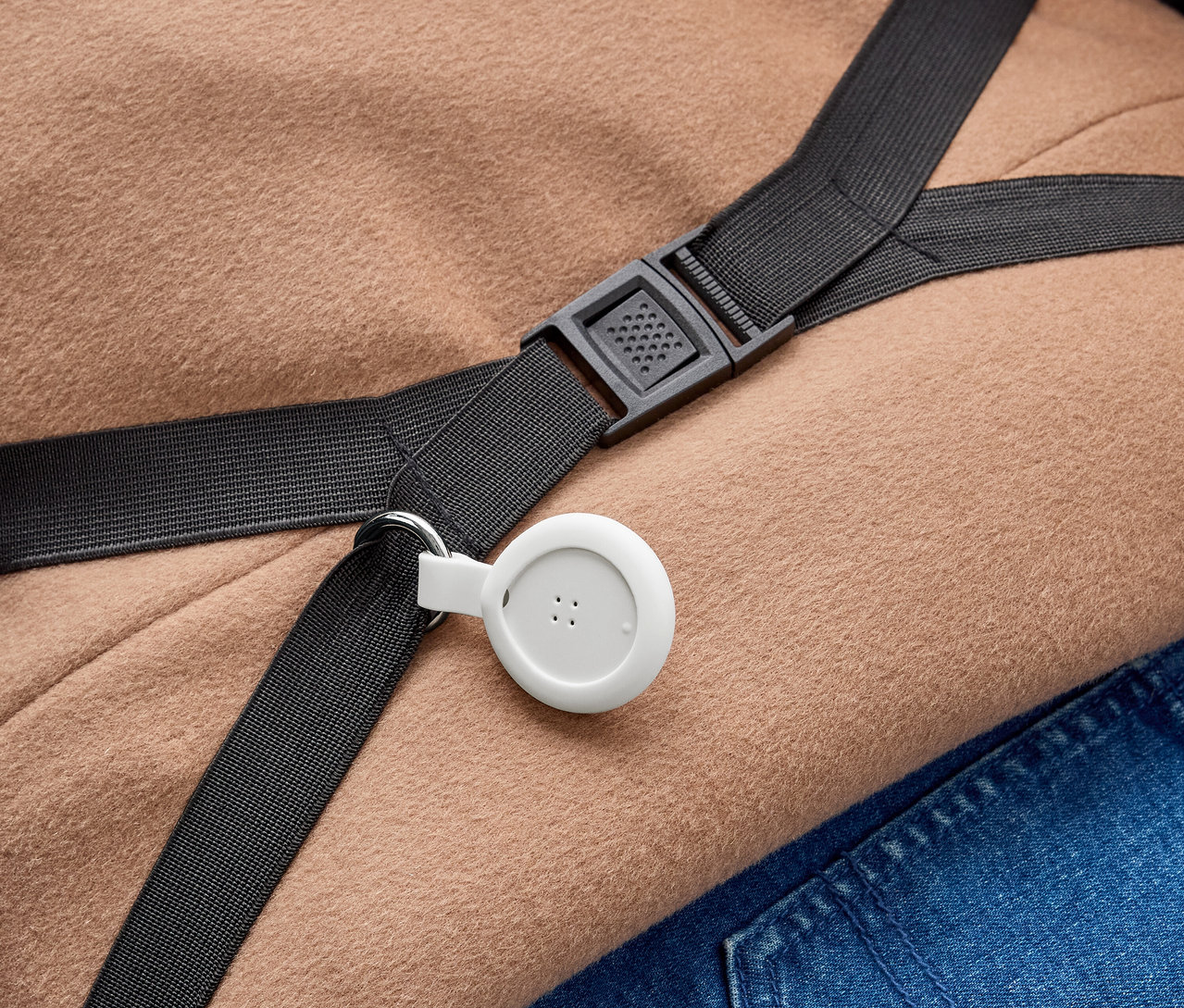 Białe, okrągłe urządzenie z trzema kropkami jest przymocowane do czarnego paska za pomocą metalowego kółka. Pasek z czarną plastikową klamrą leży na jasnobrązowym materiale i niebieskim dżinsie.