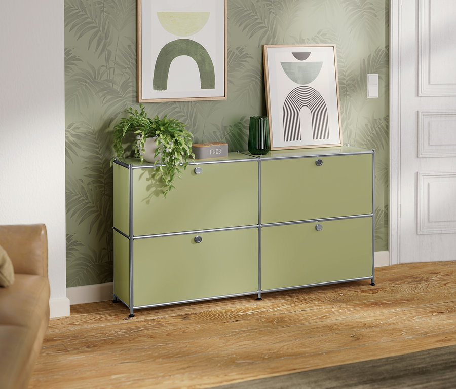 Jasnozielony kredens z czterema przegródkami stoi na tle ściany w palmy. Na kredensie stoi roślina, zegar i zielony wazon. Na ścianie wiszą dwa oprawione obrazy.