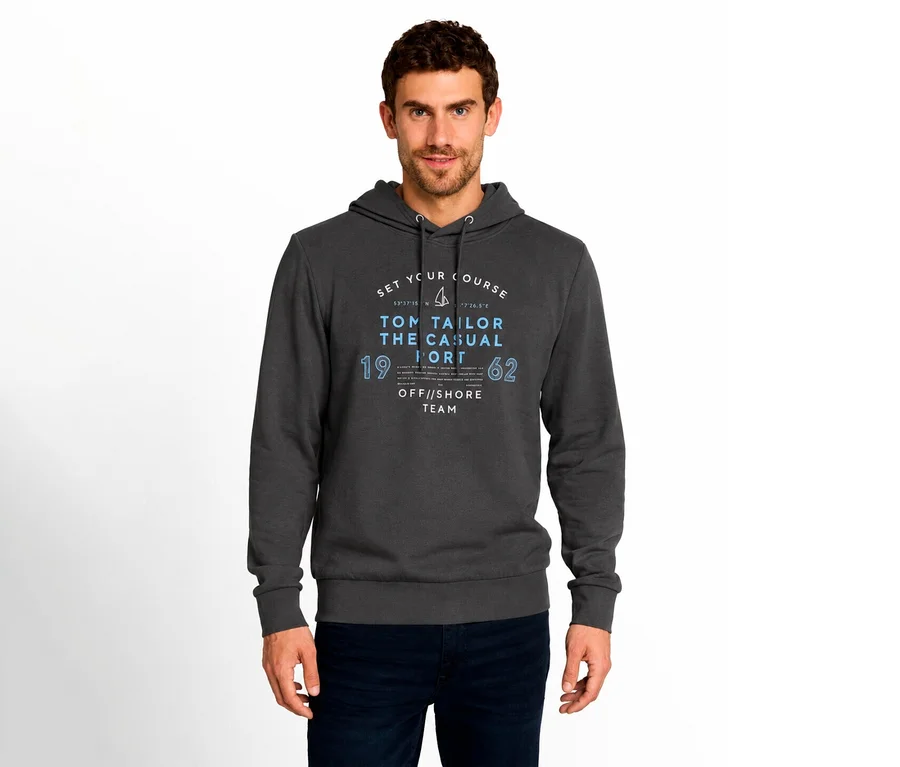 Mężczyzna w ciemnoszarej bluzie z kapturem z niebiesko-białym nadrukiem graficznym „TOM TAILOR THE CASUAL” na przodzie i ciemnoniebieskich dżinsach, stojący na białym tle.