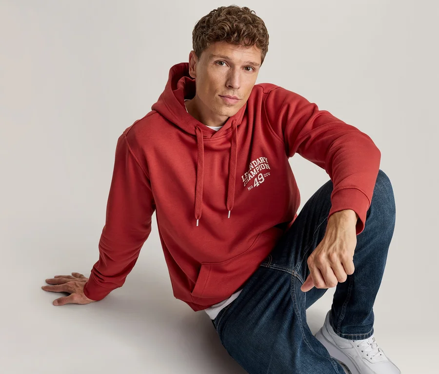 Mężczyzna siedzi na podłodze, ubrany w czerwoną bluzę z kapturem z napisem „LEGENDARY CHAMPION No. 49 DIV”, niebieskie dżinsy i białe sneakersy, patrzy w kamerę.