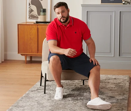 Mężczyzna siedzi na krześle ubrany w granatowe szorty chinosy, czerwoną koszulkę polo o splocie piké i buty typu sneaker.
