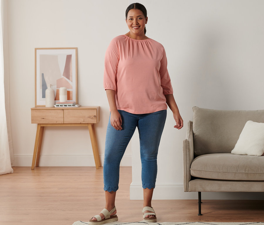 Kobieta stoi i ma na sobie sandały skórzane na platformie, tregginsy o długości 7/8 z ażurowym haftem i koralową bluzkę z rękawami o długości 3/4.