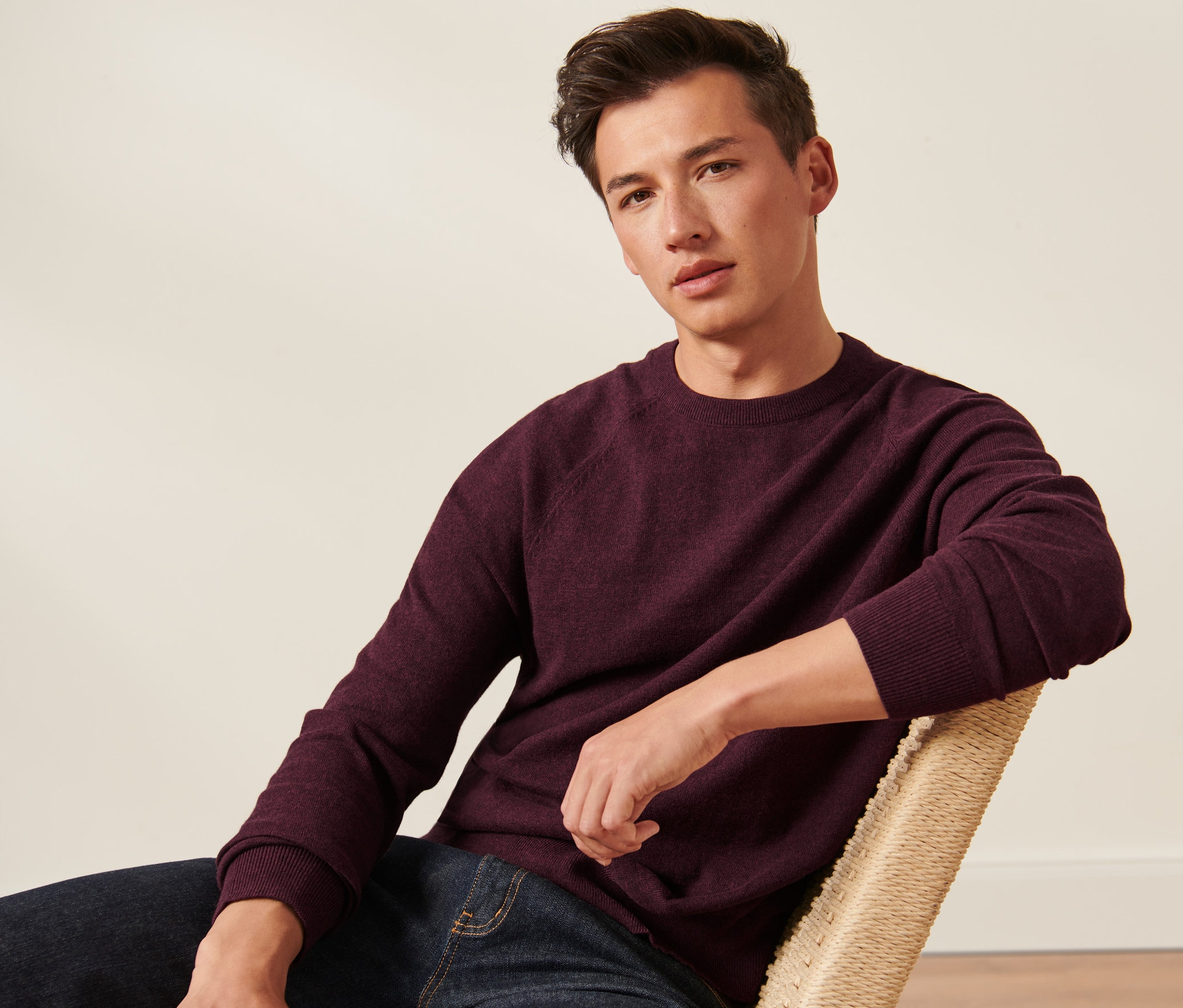 Mężczyzna siedzi na krześle i ma na sobie bordowy sweter z okrągłym wycięciem pod szyją.