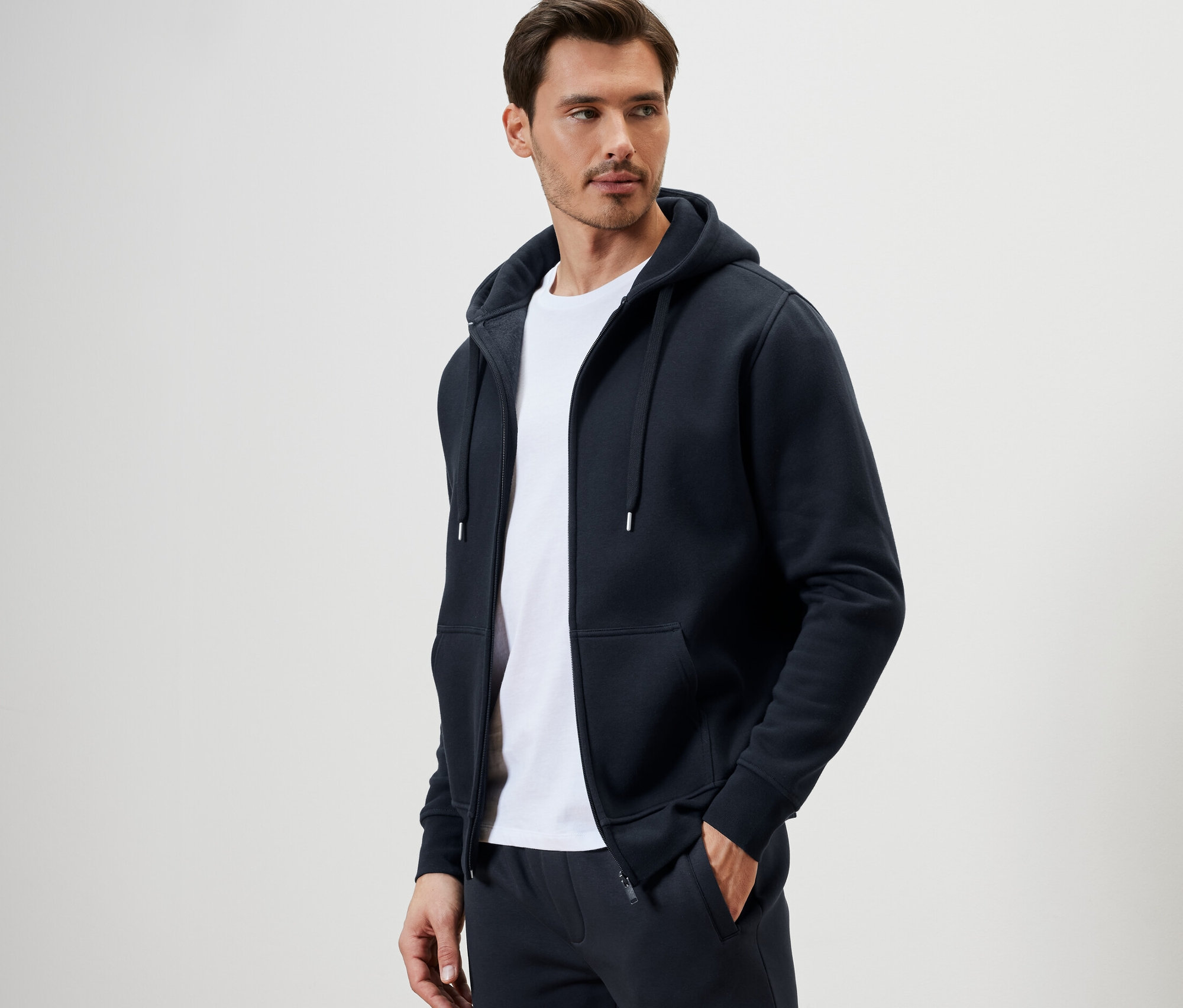 Mężczyzna ma na sobie rozpinaną bluzę z kapturem i białą koszulkę.