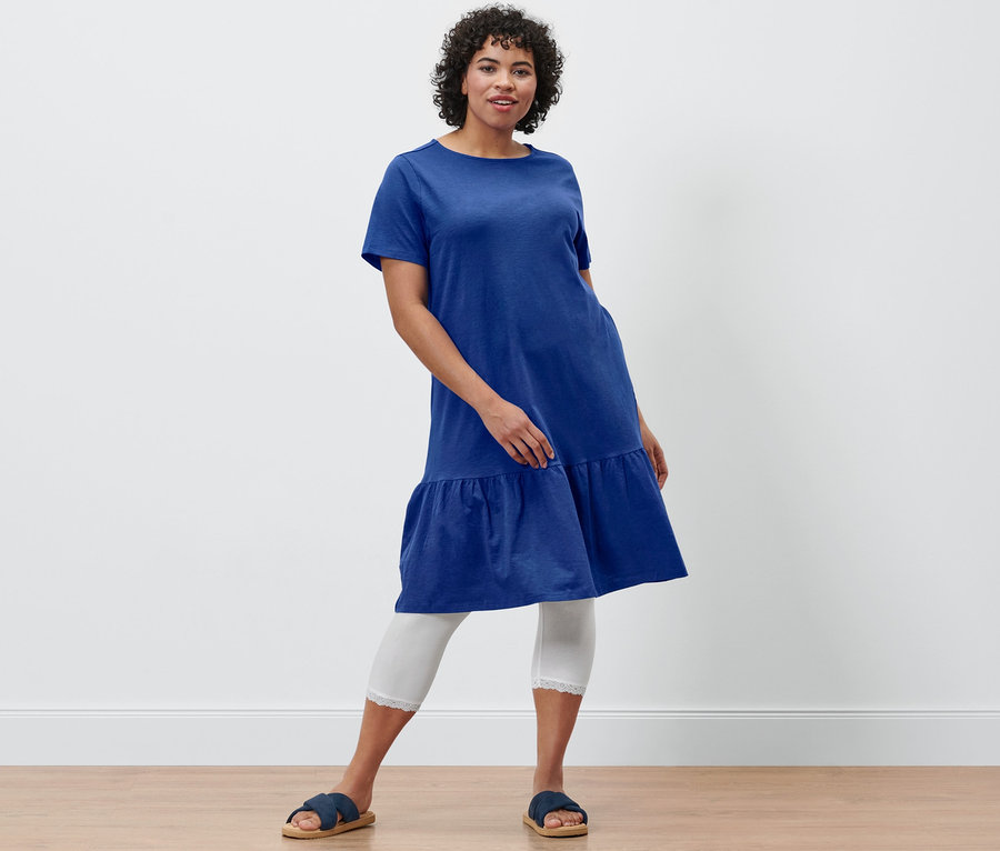 Kobieta pozuje w kobaltowej sukience dżersejowej z falbaną, białych legginsach 3/4 z koronkowym wykończeniem i wygodnych sandałach.