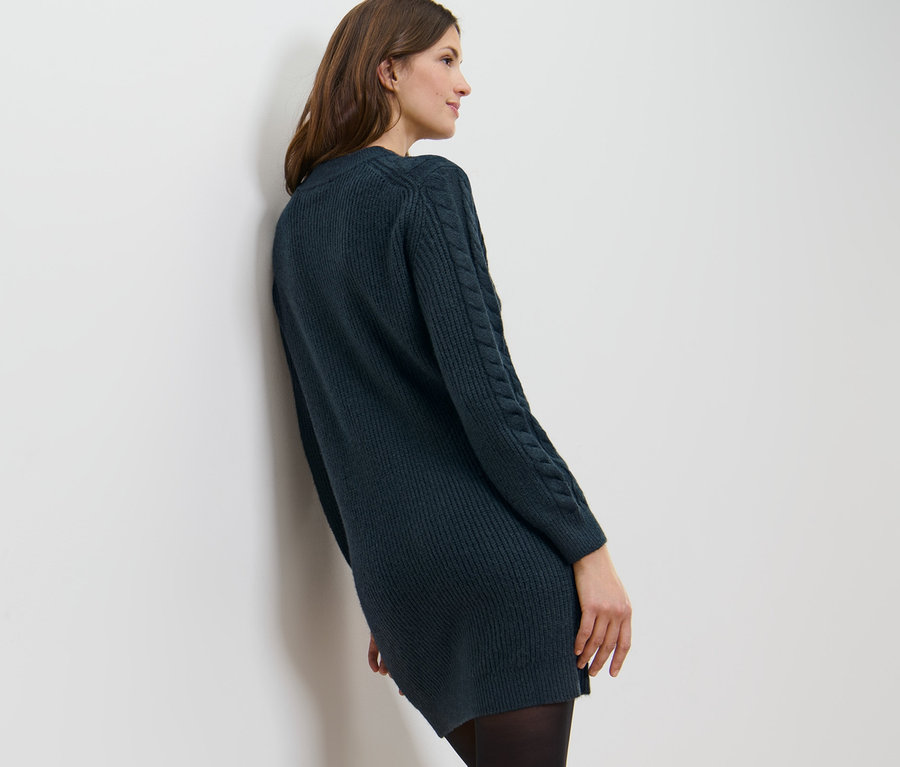 Kobieta opiera się o białą ścianę, ubrana w sukienkę z dzianiny z warkoczowym wzorem.