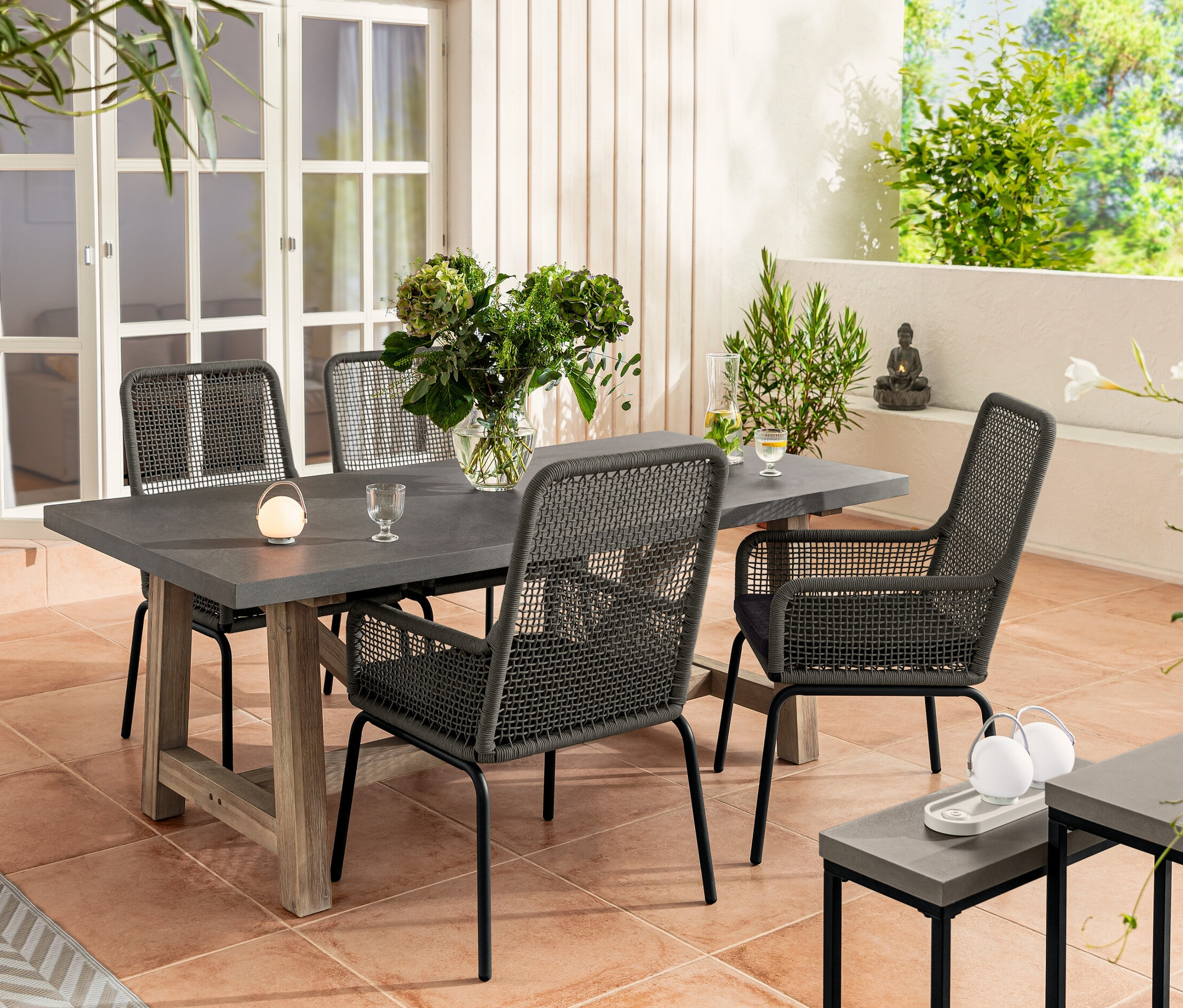 Cztery krzesła ogrodowe z wysokim oparciem z plecionką tekstylną stoją przy stole z policementowym blatem. Na stole stoi wazon z kwiatami i zestaw akumulatorowych lamp stołowych. W tle ogród i zestaw konsoli z blatem z policementu.