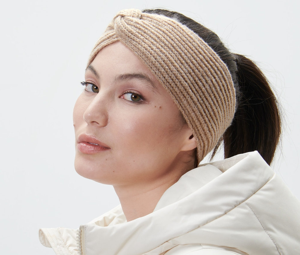 Kobieta w piaskowej opasce z dzianiny i białej kurtce patrzy przez ramię.