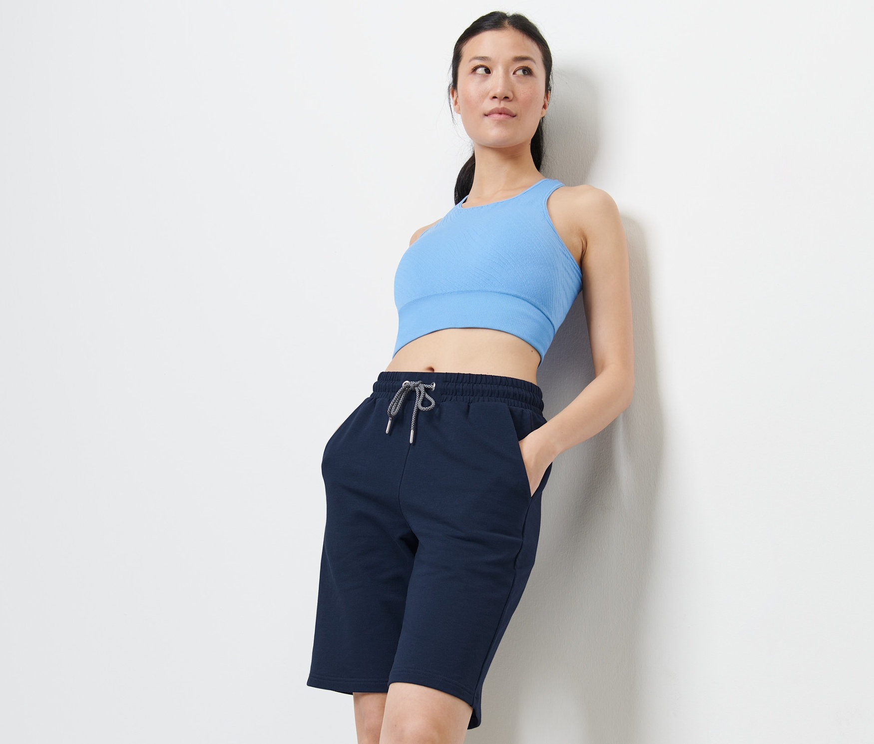 Kobieta oparta o ścianę ma na sobie niebieski top sportowy i szorty sportowe.