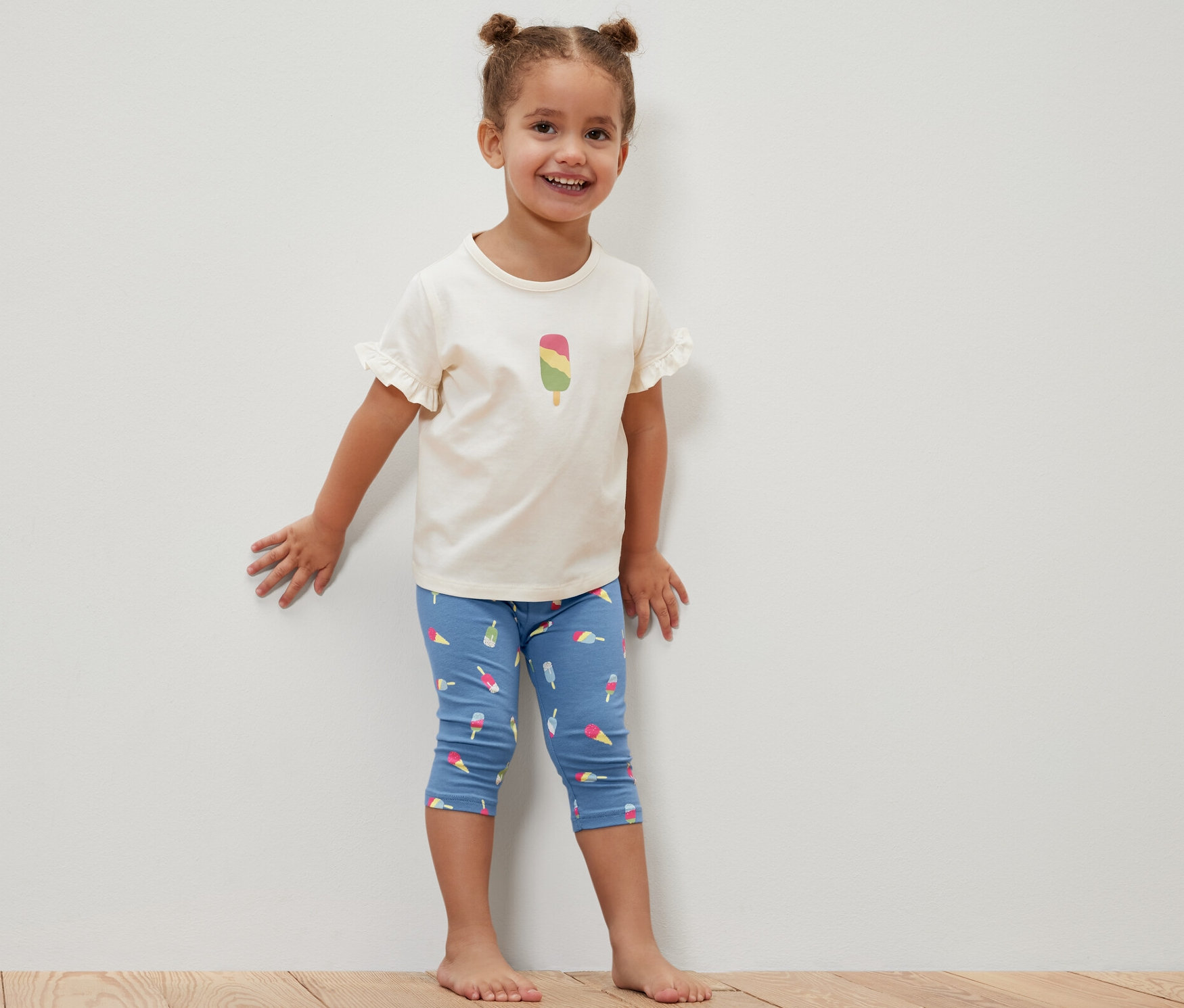 Uśmiechnięta dziewczynka stoi przy białej ścianie, ubrana w niebieskie legginsy o długości ¾ z nadrukiem lodów i białą koszulkę z rysunkiem loda. Legginsy dziecięce o długości ¾, 2 pary, niebieskie.