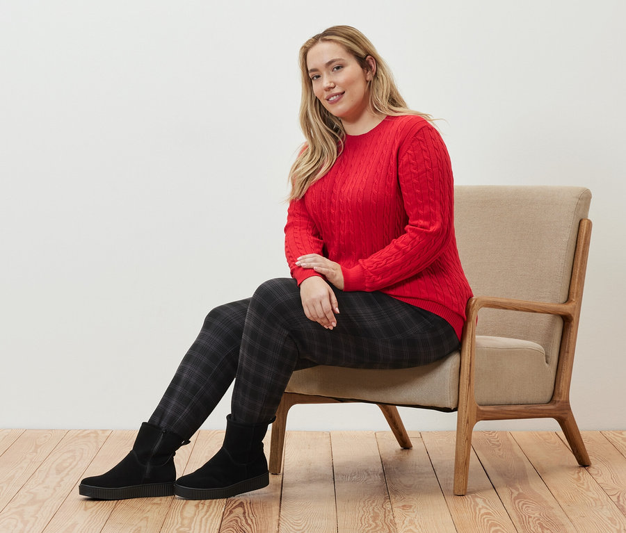 Kobieta siedzi w fotelu, ubrana w czerwony sweter z warkoczowym wzorem i spodnie joggery w kratę.