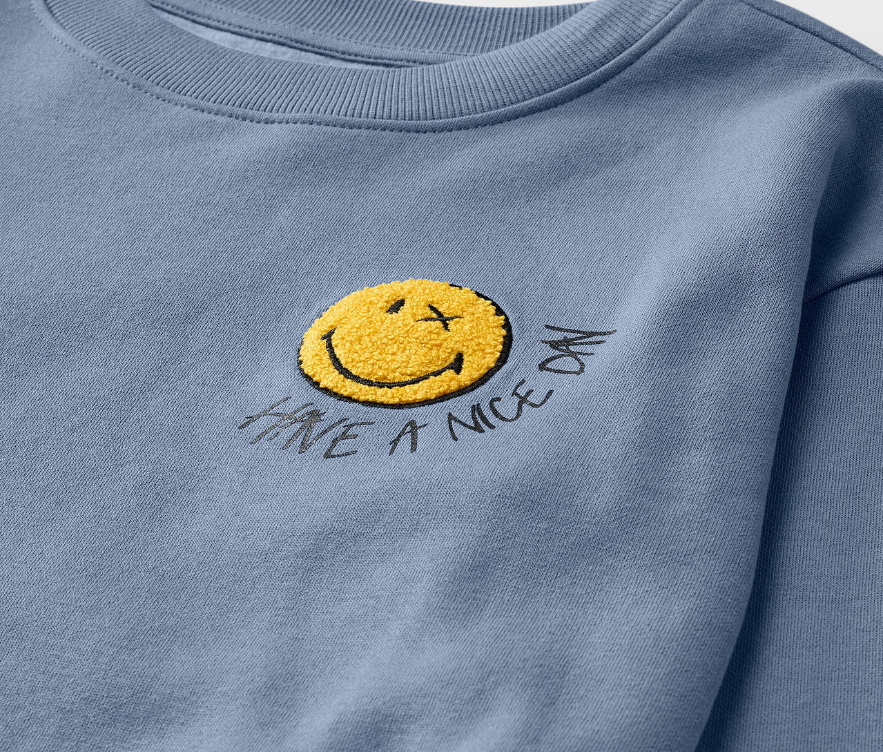 Zbliżenie na jasnoniebieską bluzę SMILEYWORLD® z żółtą buźką.