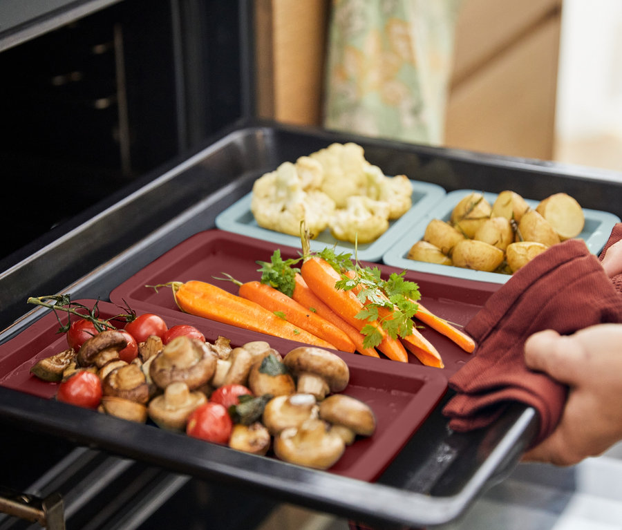 Ręka w rękawicy wyciąga z piekarnika blachę z matami do pieczenia, na których leżą warzywa.