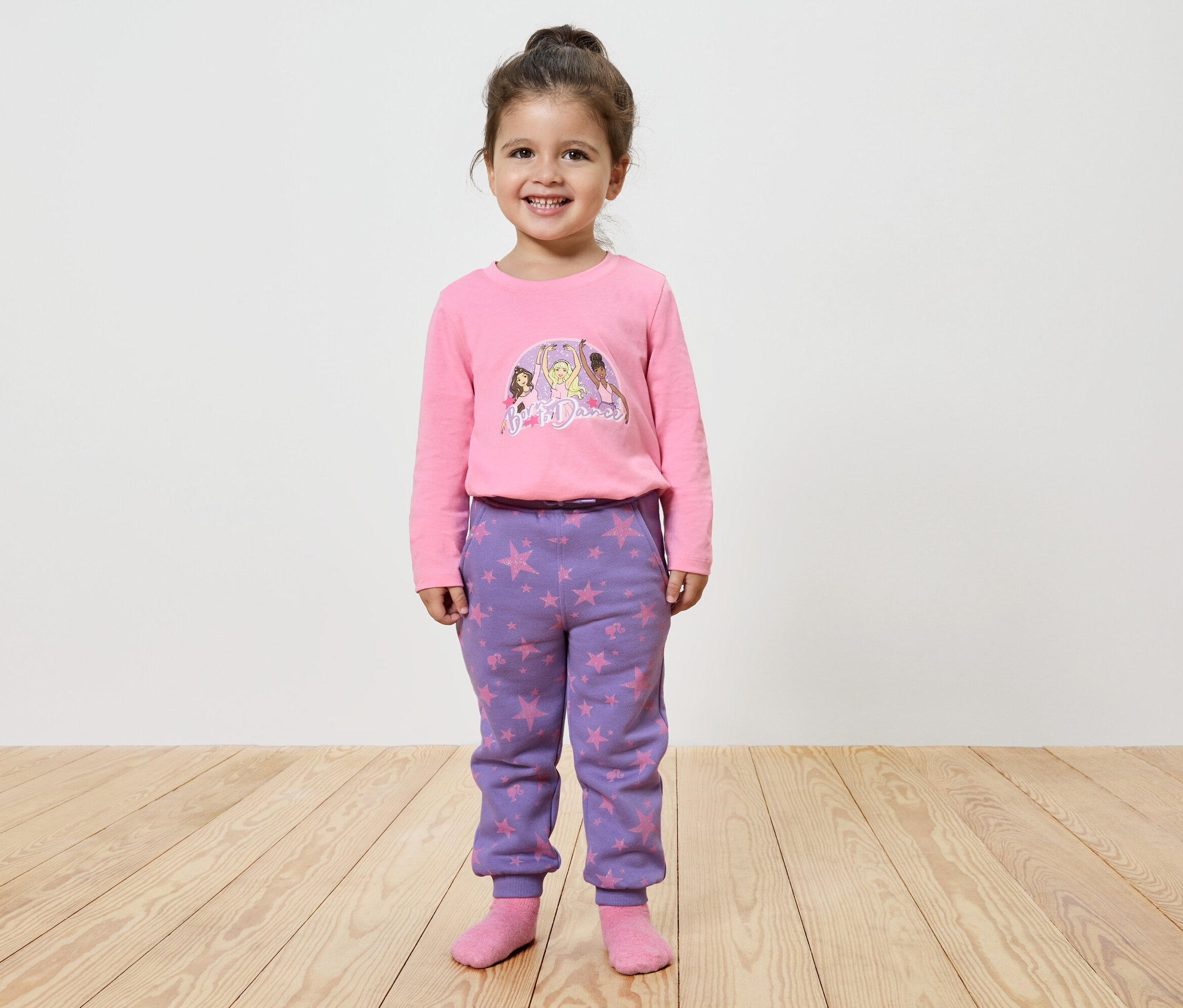 Uśmiechnięta dziewczynka pozuje w różowej koszulce z nadrukiem i fioletowych spodniach w gwiazdy.
