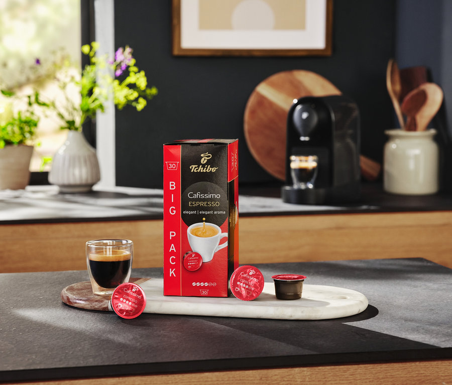 Na stole stoi opakowanie kapsułek Cafissimo Espresso, szklanka espresso i ekspres do kawy.