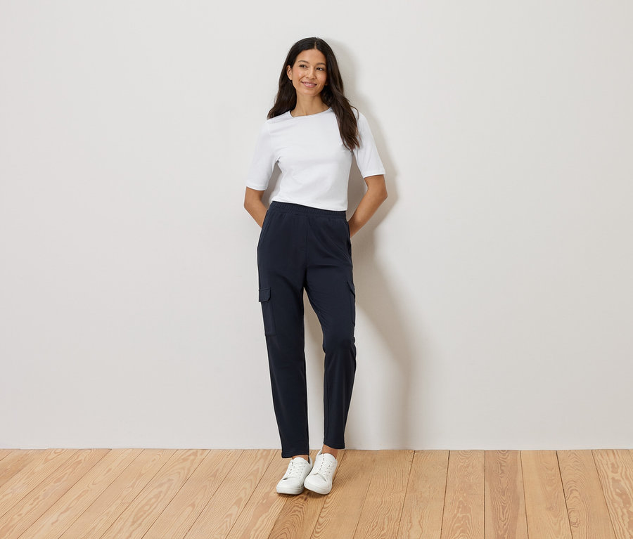 Kobieta stoi ze skrzyżowanymi rękami na tle ściany, ubrana w białą koszulkę, granatowe spodnie cargo i białe tenisówki.