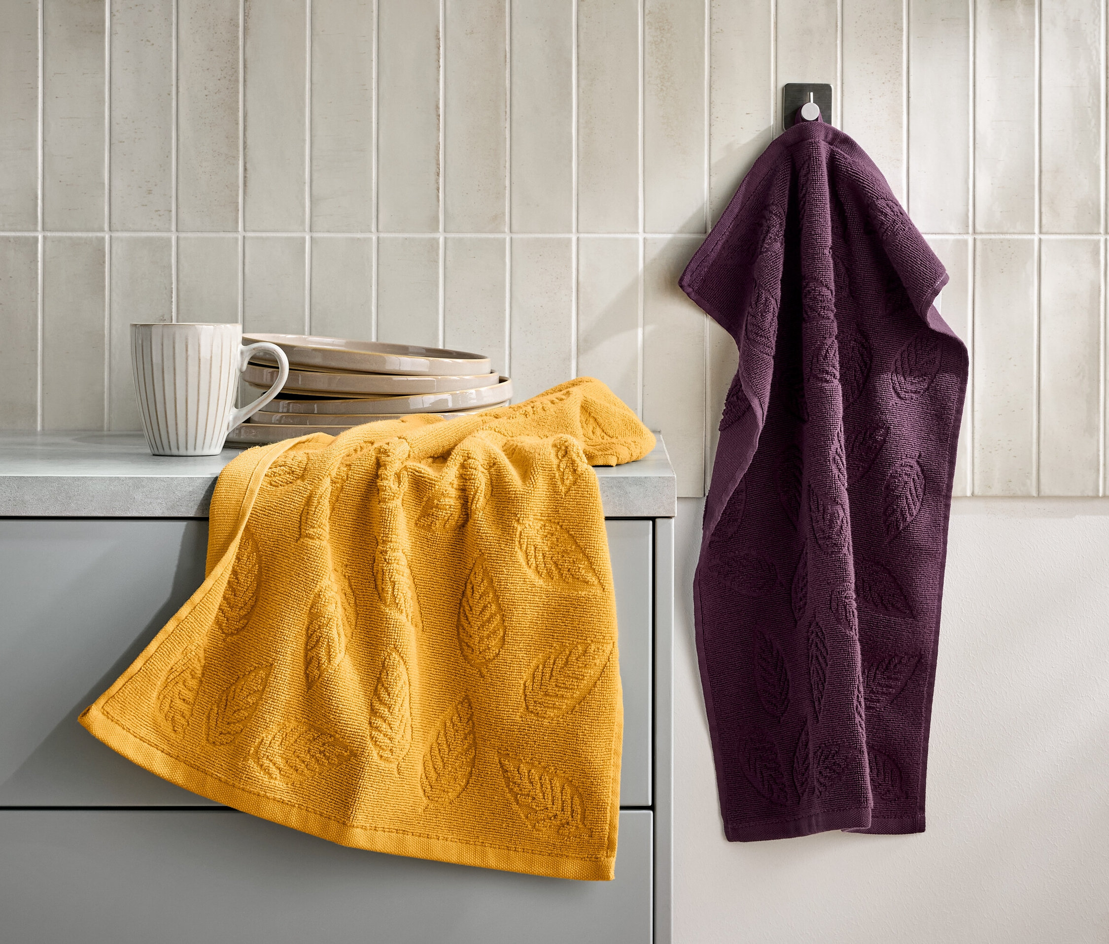Żółty ręcznik z motywem liści leży na szarej szafce obok filiżanki i talerzy. Fioletowy ręcznik wisi na haczyku na ścianie.