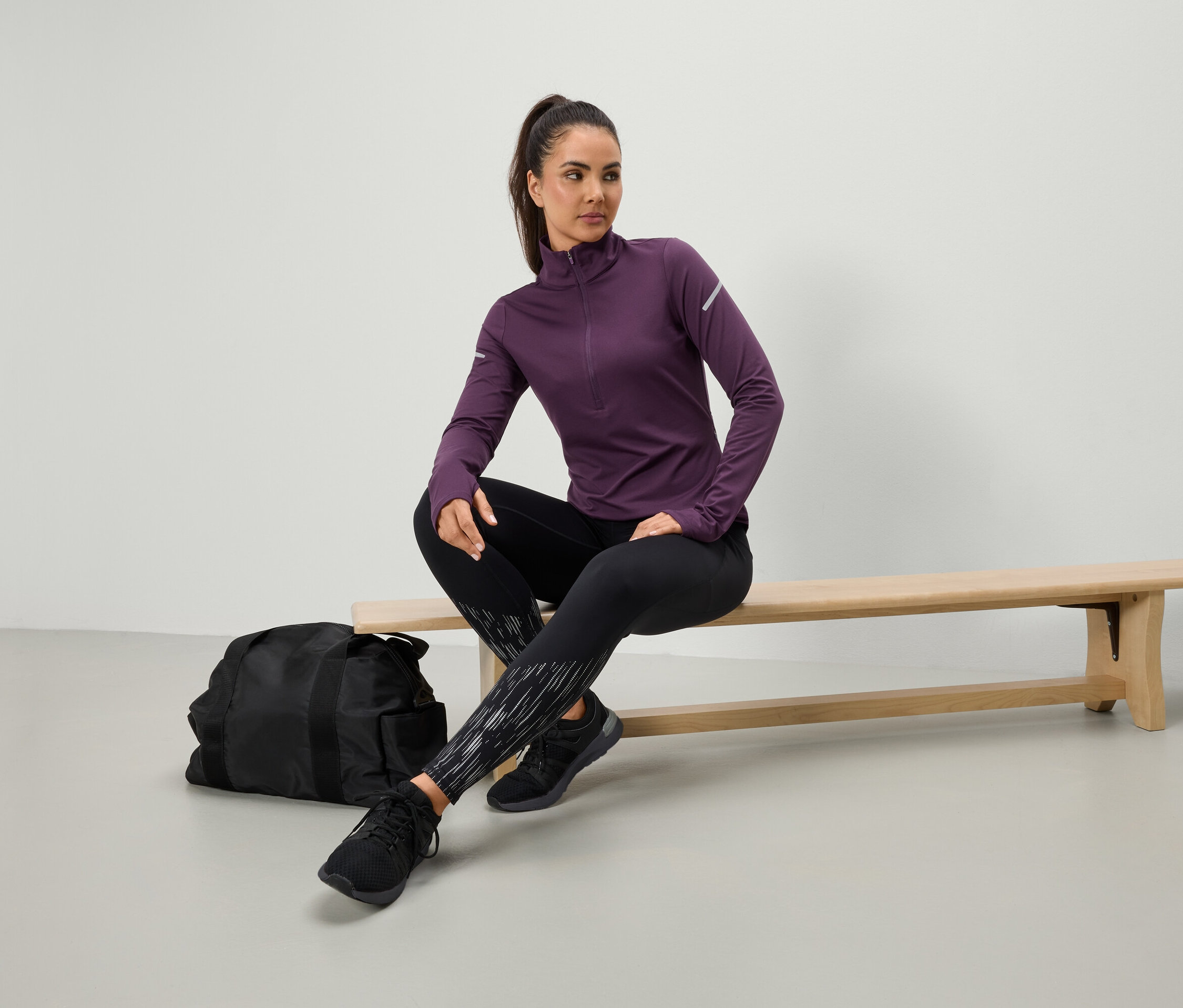 Kobieta siedzi na ławce w fioletowej koszulce sportowej i czarnych legginsach, obok niej leży czarna torba sportowa.